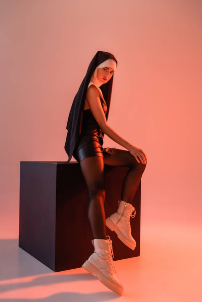 sexy nun in leather dress and boots posing on black cube on pink background - Photo, Image