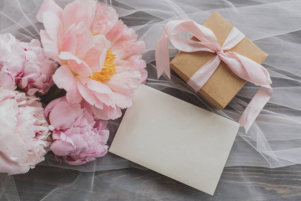 Beautiful peony bouquet, gift box and card on soft tulle fabric on dark wooden background, top view. Happy Mothers day. Greeting card template. Pink and white peonies flowers and stylish present - Photo, image