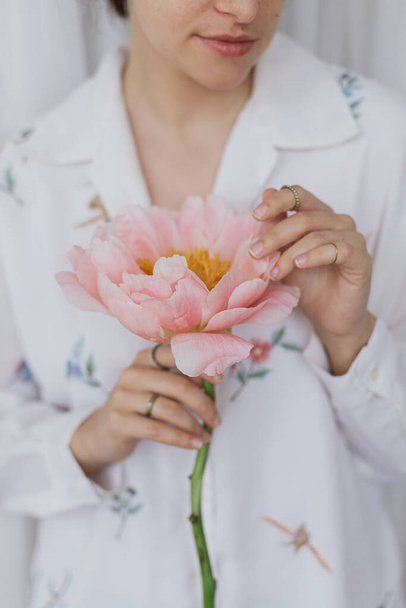 Gyönyörű elegáns nő kezében rózsaszín bazsarózsa. Fiatal nő boho virágos ingben, óvatosan fogja a nagy bazsarózsa virágot a kezében. Érzéki lágy kép. Tavaszi esztétika - Fotó, kép