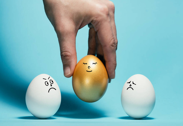 Χέρι επιλέξετε ένα χρυσό αυγό με ένα αστείο χαμογελαστό πρόσωπο. Έννοια της αποκλειστικότητας, καλύτερη επιλογή, πρόσληψη. - Φωτογραφία, εικόνα