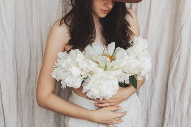 Stijlvolle sensuele vrouw met witte pioenachtige bloemen op het lichaam. Mooi jong vrouwtje met pioen boeket groeiend uit broek op beige stof achtergrond. Bloeiende lichaam positieve en bloemige geur. Zelfverzorging - Foto, afbeelding