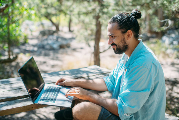 Férfi turista, aki egy laptopon dolgozik a szabadban egy kempingben. Fiatal fickó, szabadúszó, online dolgozik egy laptopon egy hegyi túrán a szabadban. A világ minden tájáról való munkavégzés képessége. - Fotó, kép