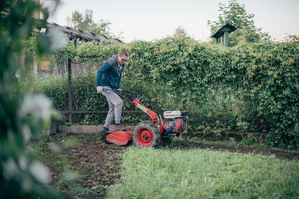 Mies työskentelee puutarhassa, kyntää maata viljelyskoneella. Nuori kaveri, jolla on käsi traktori kaivaa maata kasvimaa varten maalaistalossa - Valokuva, kuva
