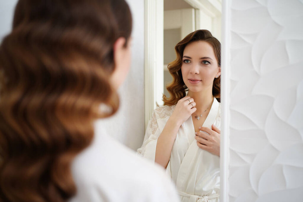 bride in a silk robe looks in the mirror and corrects the pendant around neck - Photo, image