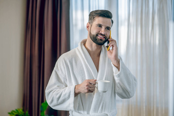 Young bearded man in a white bath robe having morning coffee and talking on the phone - Foto, afbeelding