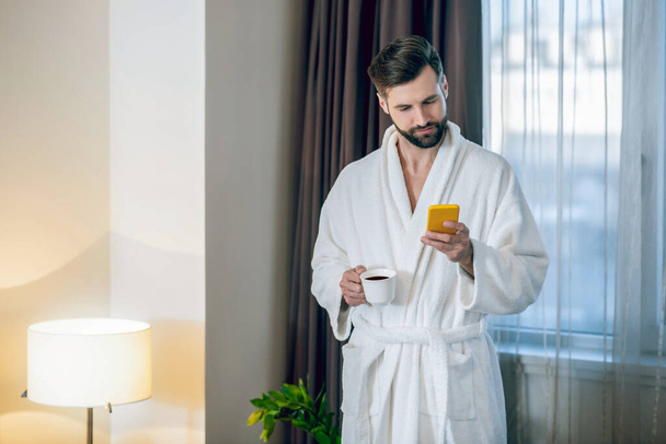 Young bearded man in a white bath robe reading a message on the phone - 写真・画像