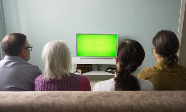 Familie kijkt TV. Groen scherm. Een gezin van vier zit thuis op de bank. Voor hen staat een groene tv..  - Foto, afbeelding