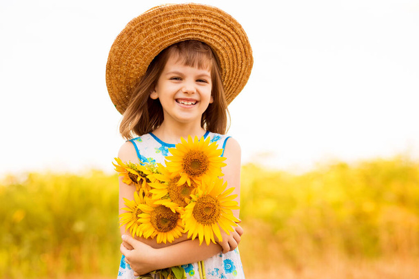 Наружный портрет очаровательной маленькой девочки в соломенной шляпе и синем платье прогуливается по полю подсолнухов, собирая и собирая букет. Летние каникулы - Фото, изображение