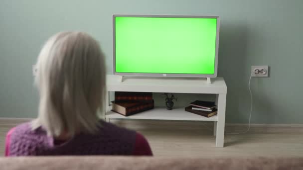 A avó está a ver televisão. Tela verde. Uma avó com cabelo grisalho senta-se no sofá em casa. Na frente dela está uma TV de tela verde. Escoamento horizontal. - Filmagem, Vídeo