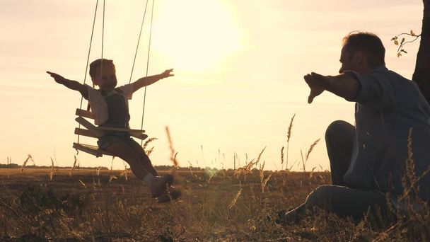 Um pai balança uma criança em um balanço em um parque de verão ao pôr-do-sol no céu, uma menina voa no ar com os braços estendidos e sonhando em voar, uma criança joga um avião com seu pai, uma filha - Foto, Imagem