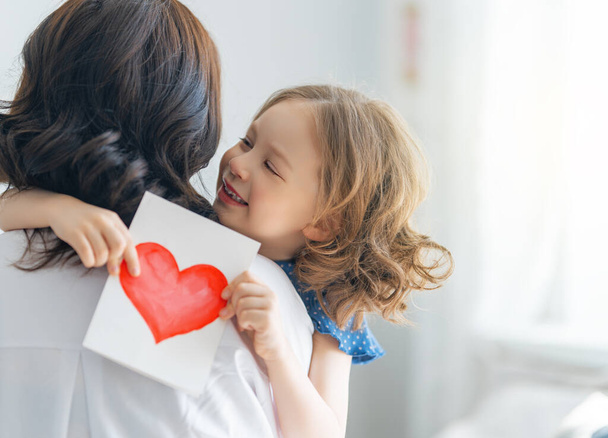 Happy mother's day! Child daughter is congratulating mom and giving her postcard. Mum and girl smiling and hugging. Family holiday and togetherness. - Фото, зображення