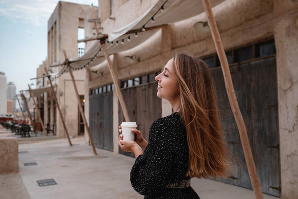 Donna felice viaggiatrice vestita di nero che cammina per le strade di una vecchia città araba o villaggio nel mezzo del deserto. caffè in una tazza bianca, gustando caffè arabo tradizionale - Foto, immagini