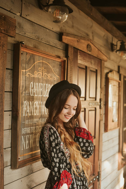 Meisje in het Wilde Westen, in het Westerse huis. Meisje met hoed met lange cerly haar glimlach. Mooi mooi meisje met een zwarte hoed. Ongelooflijke reis, reizen. Vrouw op het terras in rode jurk pouse. - Foto, afbeelding