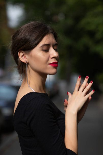 Mulher morena elegante elegante com lábios vermelhos e manicure usa vestido preto posando sob a rua com luz solar - Foto, Imagem