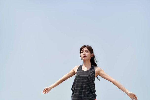 joven asiático mujer en sportswear de pie al aire libre contra azul cielo con abierto brazos - Foto, Imagen
