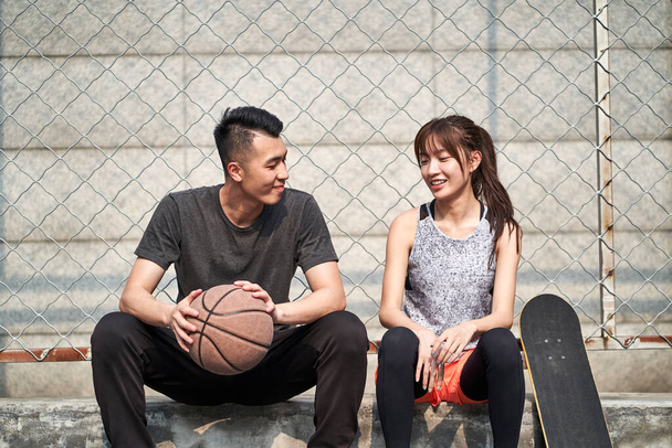 若いアジア系成人男性バスケットボール選手と女子スケートボーダーがコートサイドでチャット - 写真・画像
