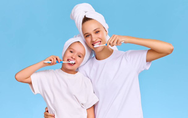 Entzückte junge Frau und Mädchen mit Handtüchern auf nassem Haar, die sich umarmen und lächelnd in die Kamera schauen, während sie sich vor blauem Hintergrund die Zähne putzen - Foto, Bild