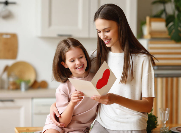 Happy young woman hugging girl and reading greeting card with heart during Mother day celebration in kitchen at home - Foto, imagen
