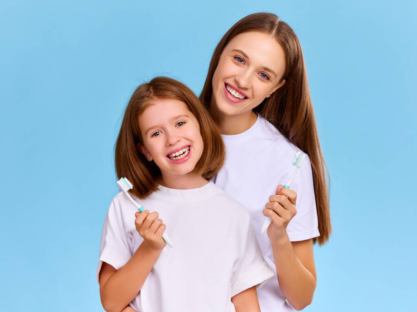 Optimistic family: woman and girl in white t shirts smiling and looking at camera while brushing teeth together against blue background - Photo, Image