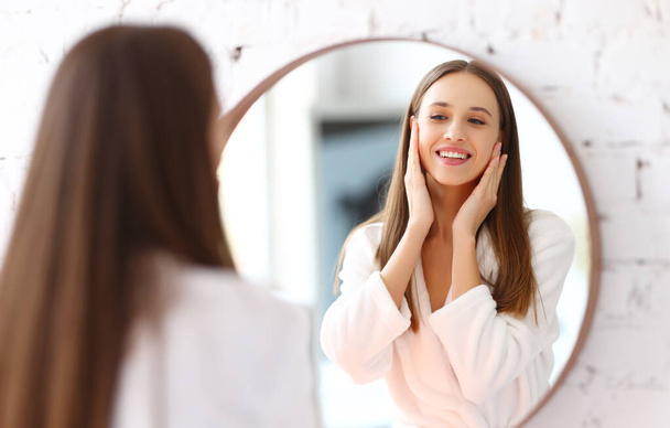 Glückliche junge Frau im Bademantel, die kosmetische Creme auf das Gesicht aufträgt und die Weichheit der Haut während einer Wellness-Beauty-Session zu Hause genießt - Foto, Bild