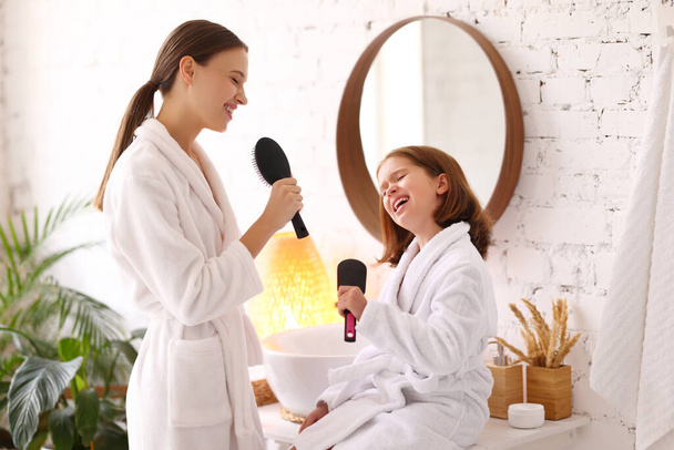 Side view of young woman and little daughter in white bathrobes holding hairbrushes in hands as microphones and singing together while enjoying beauty procedure at home - Photo, Image