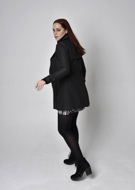 Dlouhý portrét rudovlasé dívky v černém kabátě, tartanové sukni a botách. Stojící póza se zády ke kameře na pozadí studia. - Fotografie, Obrázek