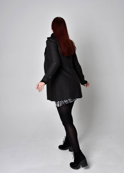 Ganzkörperporträt eines rothaarigen Mädchens mit schwarzem Mantel, Schottenrock und Stiefeln. Vor Studiohintergrund mit dem Rücken zur Kamera posieren. - Foto, Bild
