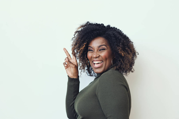 Side view of cheerful curly haired woman showing peace sign against white background - Photo, image