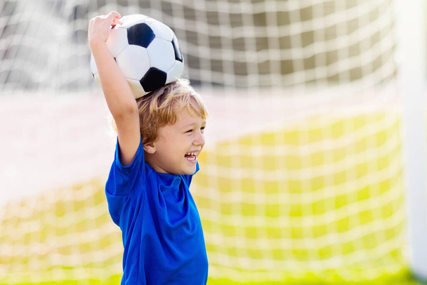 Τα παιδιά παίζουν ποδόσφαιρο σε υπαίθριο γήπεδο. Τα παιδιά σκοράρουν ένα γκολ κατά τη διάρκεια του αγώνα ποδοσφαίρου. Μικρό αγόρι κλωτσάει μπάλα. Σχολική αθλητική λέσχη. Κατάρτιση για νέους παίκτες. - Φωτογραφία, εικόνα