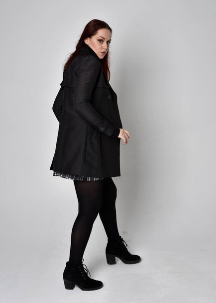 Портрет повної довжини рудої волохатої дівчини в чорному светрі з черепашками, спідниці та чоботях. Стоїть поза спиною до камери на фоні студії
. - Фото, зображення