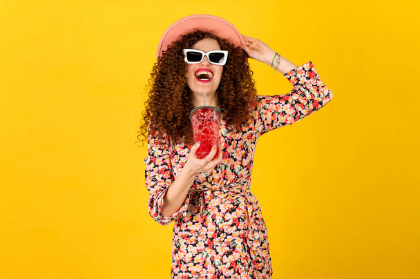 Dívka v letním oblečení na sobě bílé trendy sluneční brýle a těší při pití červeného koktejlu. Pózování na žlutém pozadí. - Fotografie, Obrázek