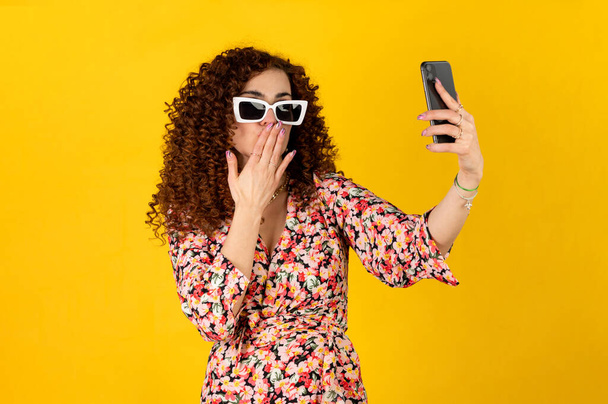 Girl in summer outfit with curly hair using smartphone sending kisses while looking at the phone camera. Isolated on yellow background. - Фото, изображение