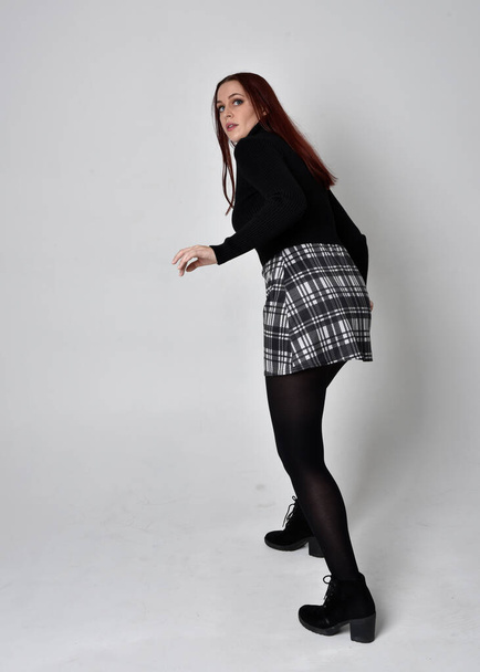 Retrato de cuerpo entero de una chica pelirroja con suéter de cuello alto negro, falda a cuadros y botas. Posar de pie con la espalda a la cámara sobre un fondo de estudio. - Foto, imagen