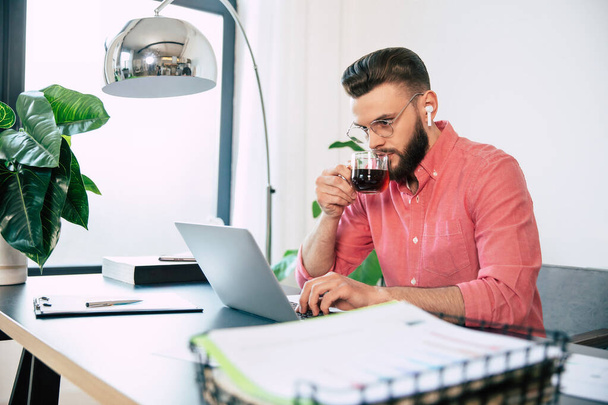 Confident successful handsome bearded man in eyeglasses and smart casual wear is working on the laptop at modern co-working or home office - 写真・画像