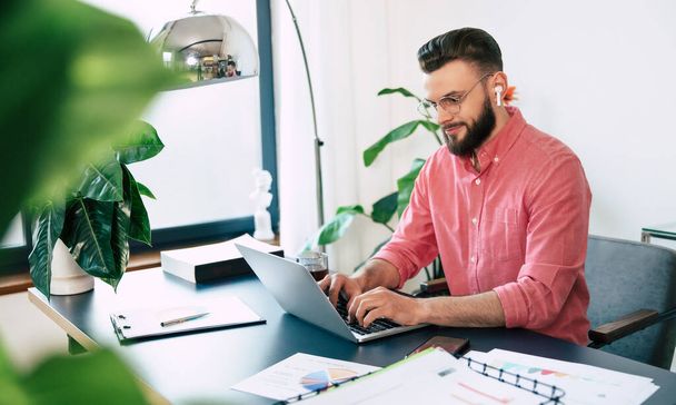 Confident successful handsome bearded man in eyeglasses and smart casual wear is working on the laptop at modern co-working or home office - Фото, зображення