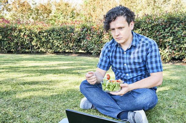 Junger Mann, der in einer Pause im Gras sitzt und einen frischen Salat isst, während er arbeitet oder am Laptop lernt. Konzepte für Arbeit im Freien und gesundes Leben und Essen. - Foto, Bild
