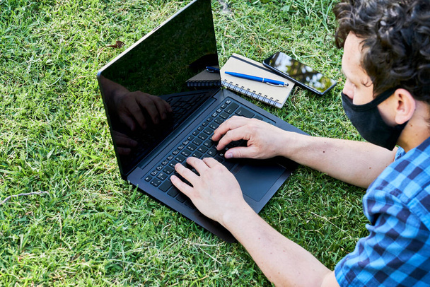 Young man wearing a cloth face mask working or studying using a laptop outdoors. Concepts of remote working and protection from coronavirus, during the Covid pandemic. - Photo, Image