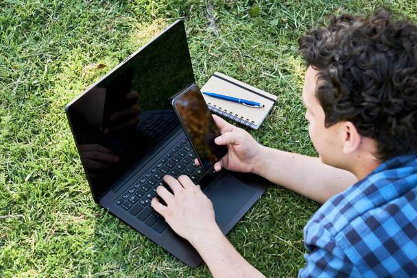 Jongeman met behulp van een smartphone en een laptop buiten, op het gras. Begrippen telewerken en studeren. - Foto, afbeelding