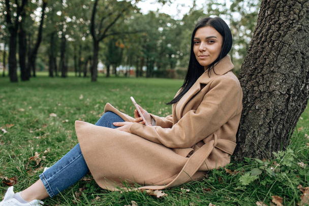 Beautiful brunette in a brown coat smiling while sitting under a tree in a green park - Valokuva, kuva