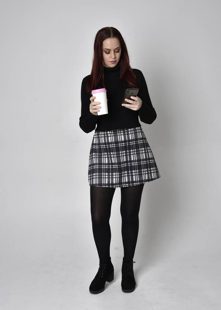 Portret rudej dziewczyny w czarnym swetrze, spódnicy w kratę i butach. Stanowisko stojące na tle studia. - Zdjęcie, obraz