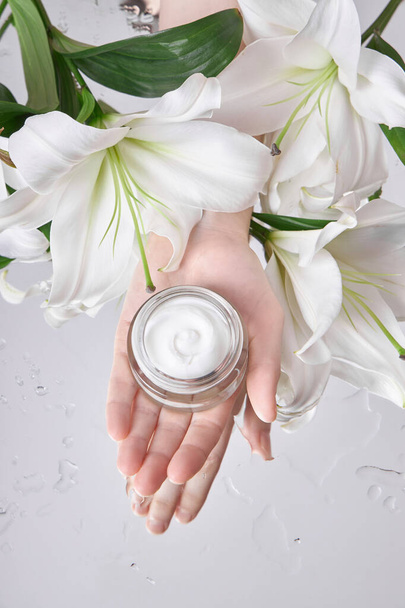Beauty jar cream hand cosmetic woman with lily flowers lie on table, jar cream product in hand woman on wet flower mirror moistening. Natural flower jar cream cosmetics for hand skin care - Photo, image