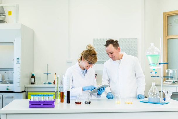 Male and female scientists working with tablet in pharmaceutical laboratory. They focus on CBD and CBDa extracts in oil and crystal form. Glass tubes, erlenmeyer flasks and separatory funnel are part of laboratory equipment on table. Healthcare pharm - Photo, Image