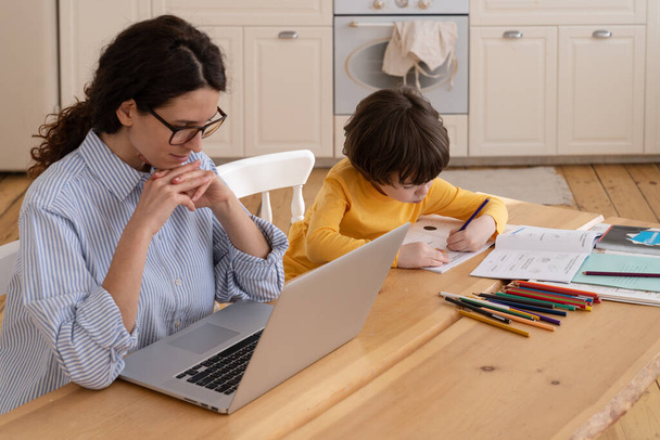 Skoncentrowany casual businesswoman czytać e-maile biznesowe na laptopie w pobliżu mały syn z biura - Zdjęcie, obraz