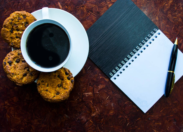 Egy csésze kávé kerek sütit egy csészealjon, egy jegyzettömb mellett egy tollal. A munka reggelivel kezdődik.. - Fotó, kép