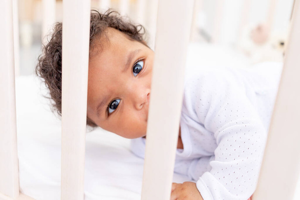 cute African-American little baby in a white bed for sleeping looks over the side - Fotoğraf, Görsel