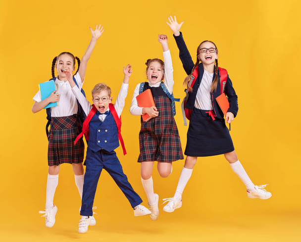 Grupa zabawnych podekscytowanych kolegów z klasy dziewcząt i chłopców w szkolnych strojach z plecakami i copybookami bawiących się i skaczących na żółtym tle - Zdjęcie, obraz