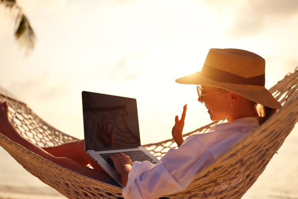 Tener una conversación en línea. Joven feliz mujer exitosa con sombrero de paja ondeando y sonriendo mientras que tiene videollamada en el ordenador portátil digital, relajarse en la hamaca en la playa de arena tropical en el día soleado - Foto, Imagen