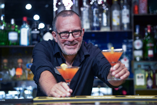 Elderly bartender with eyeglasses serving cocktail at the bar. Evening time - Photo, Image