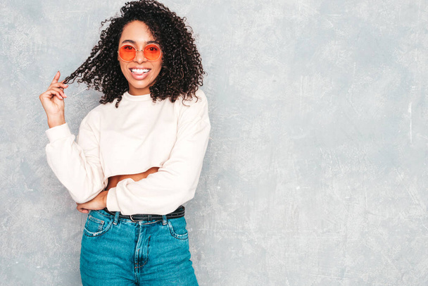 Beautiful black woman with afro curls hairstyle.Smiling model in white trendy jeans clothes and sunglasses. Sexy carefree female posing near gray wall in studio. Tanned and cheerful  - 写真・画像