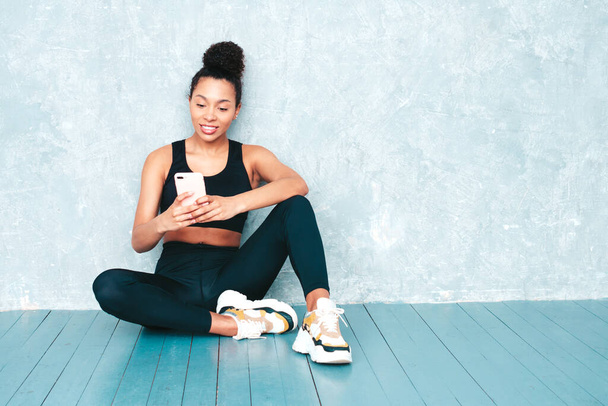 Fitness smiling black woman in sports clothing with afro curls hairstyle.She wearing sportswear. Beautiful model with perfect tanned body.Female sitting in studio. Using apps at smartphone - Valokuva, kuva